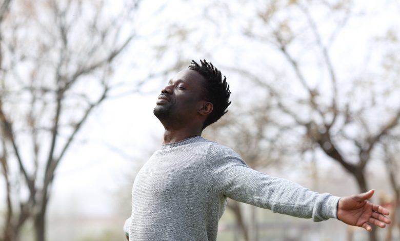 Black man breathing fresh air outdoors. Arms spread wearing a gray long sleeved shirt.