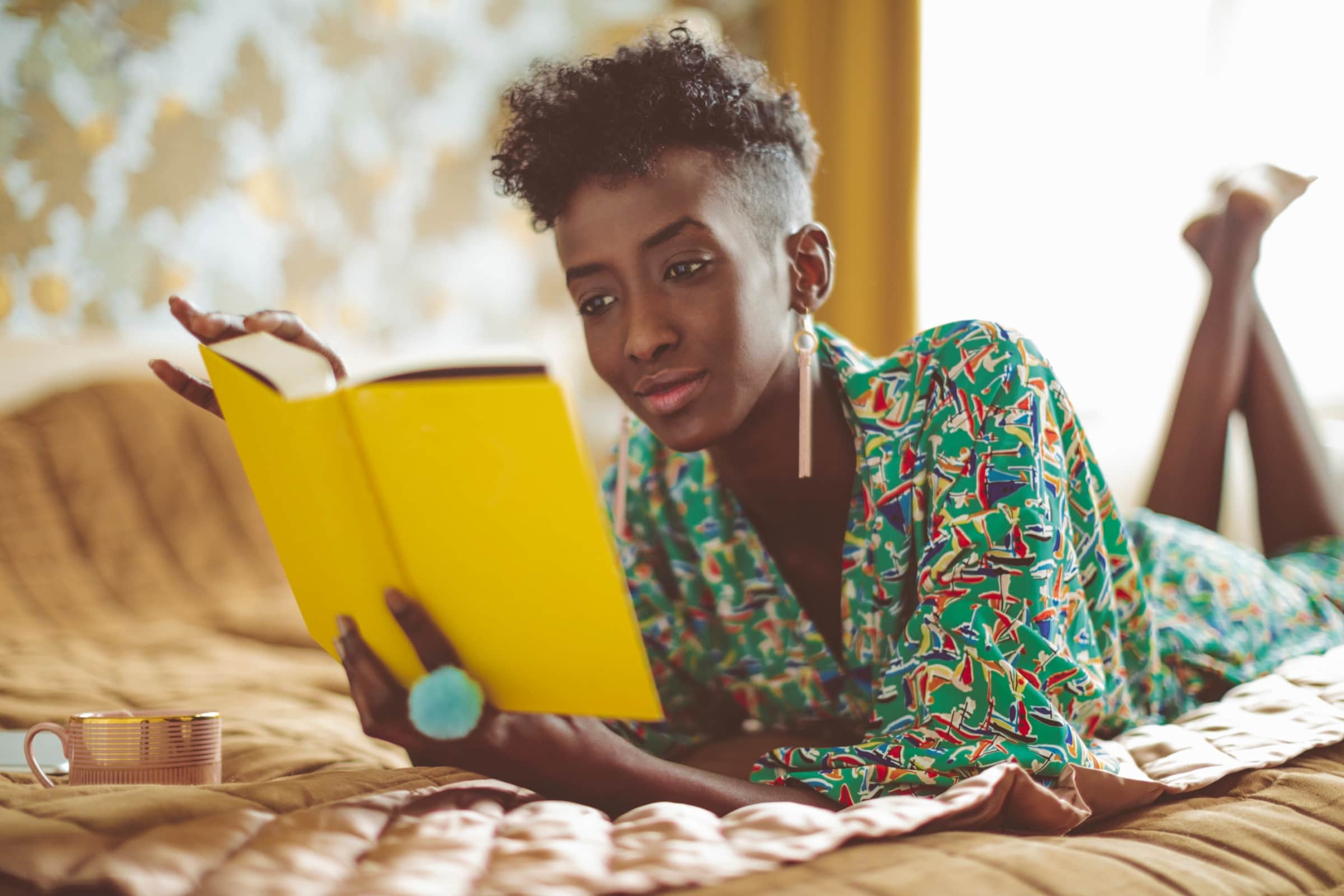 Dark-skinned Black women lying on bed reading a hard back yellow book.